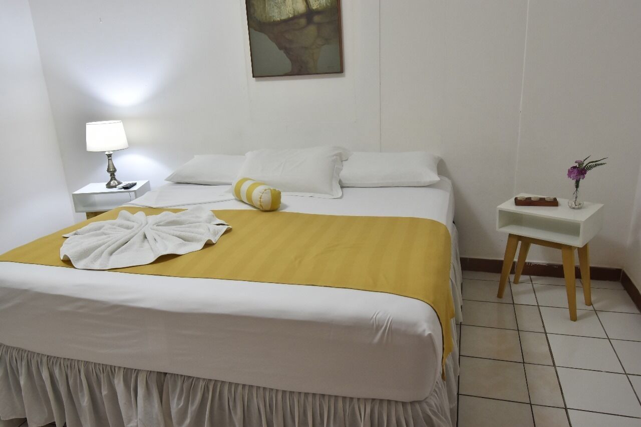 Una cama bien hecha con una manta amarilla, almohadas blancas y una toalla blanca sobre la cama. Hay una mesita de noche pequeña con una lámpara y un florero con flores.