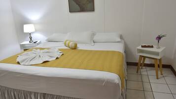 Un lit bien fait, recouvert d’une couverture jaune, avec des oreillers blancs et une serviette blanche posée sur le lit. On y trouve une petite table de chevet avec une lampe et un vase rempli de fleurs.