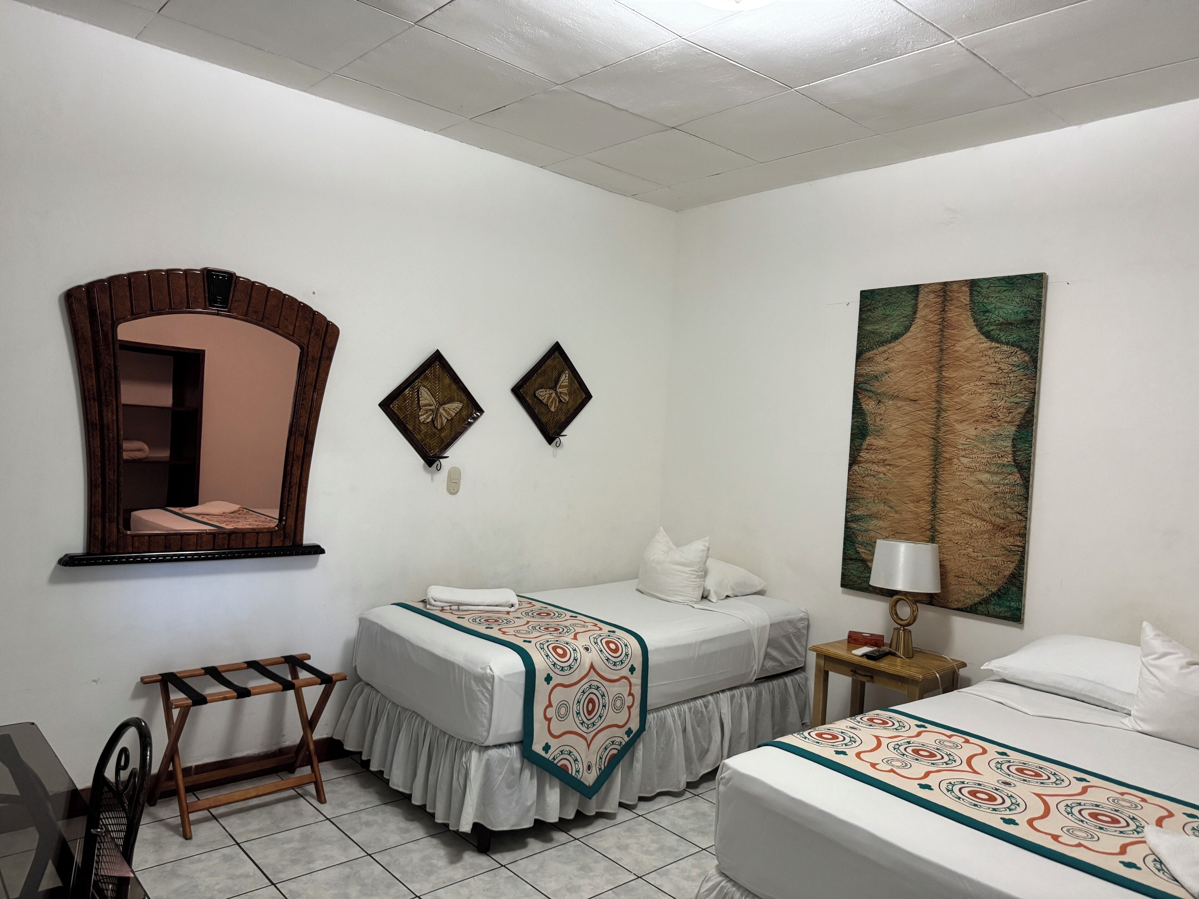 A hotel room with two single beds, a wooden chair, a small table with a lamp, and a large painting on the wall.