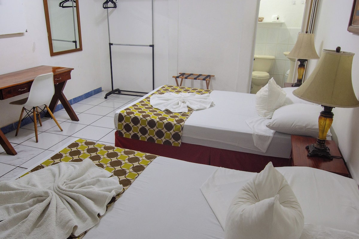 Habitación de hotel con dos camas, un escritorio, una silla, un espejo y un baño visible a través de una puerta abierta.