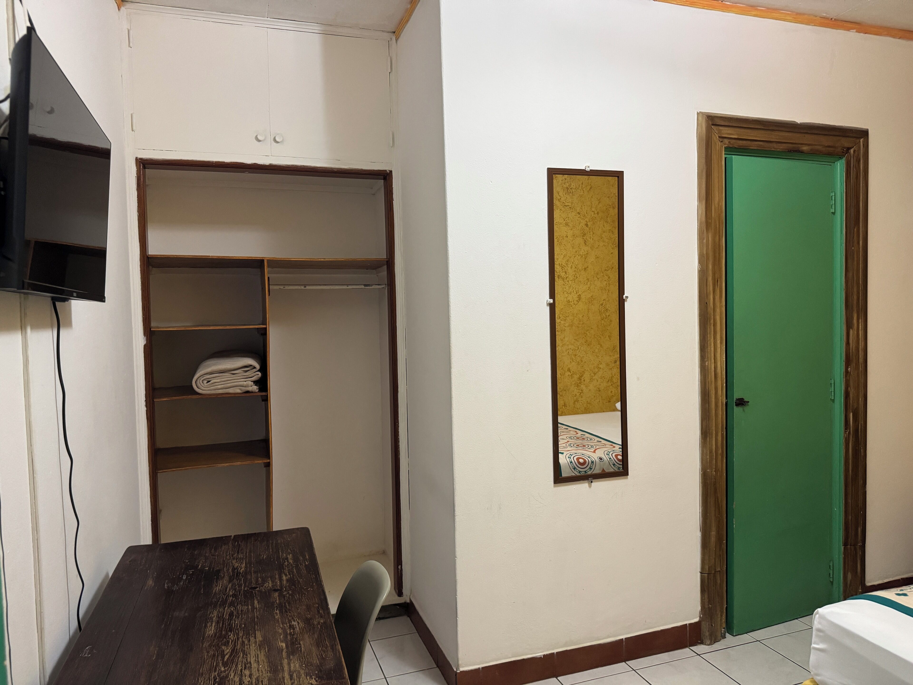 A room with a wooden table, a chair, a wardrobe, a green door, and a small mirror on the wall.