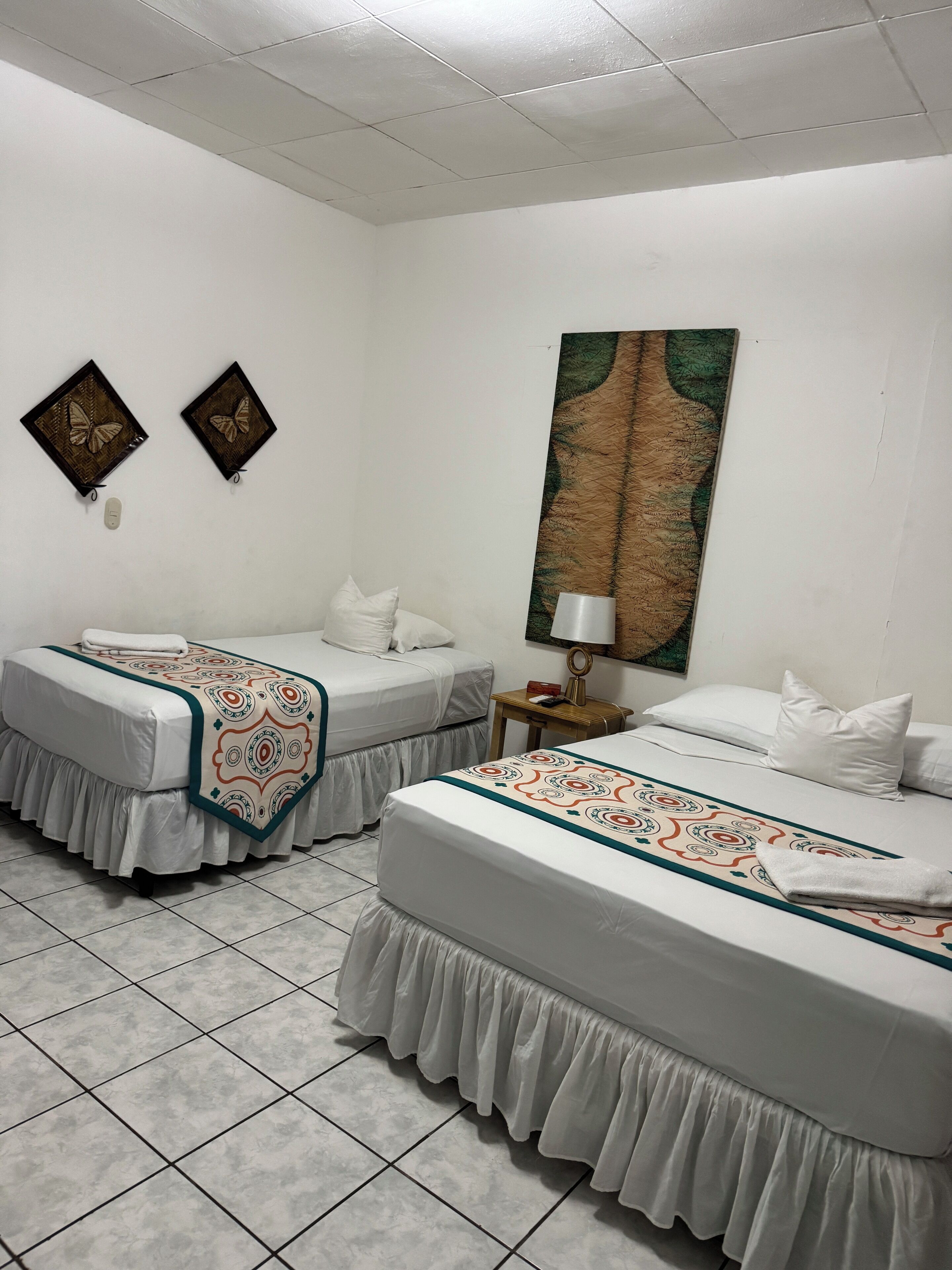 A hotel room with two single beds, a large painting on the wall, and a small table with a lamp.