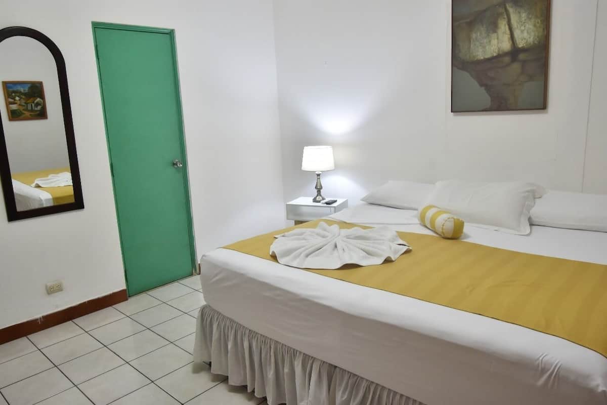 Un cuarto de hotel con cama, una mesita de noche con lámpara, una puerta verde y un cuadro en la pared.