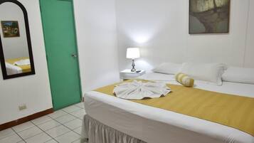 A hotel room with a bed, a nightstand with a lamp, a green door, and a painting on the wall.