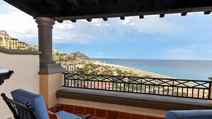Un balcon avec deux chaises bleues, une petite table sur laquelle se trouvent des fruits, et une vue sur la plage et le paysage urbain.
