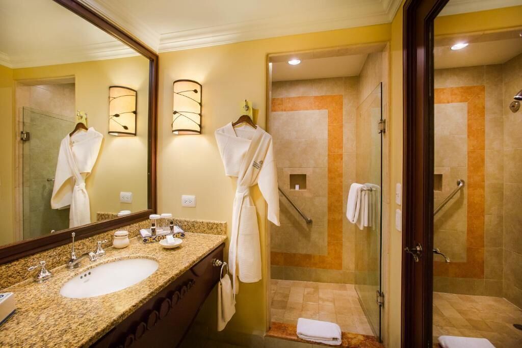 A hotel bathroom with a large mirror, a granite countertop sink, a glass shower enclosure, and two white robes hanging on hooks.