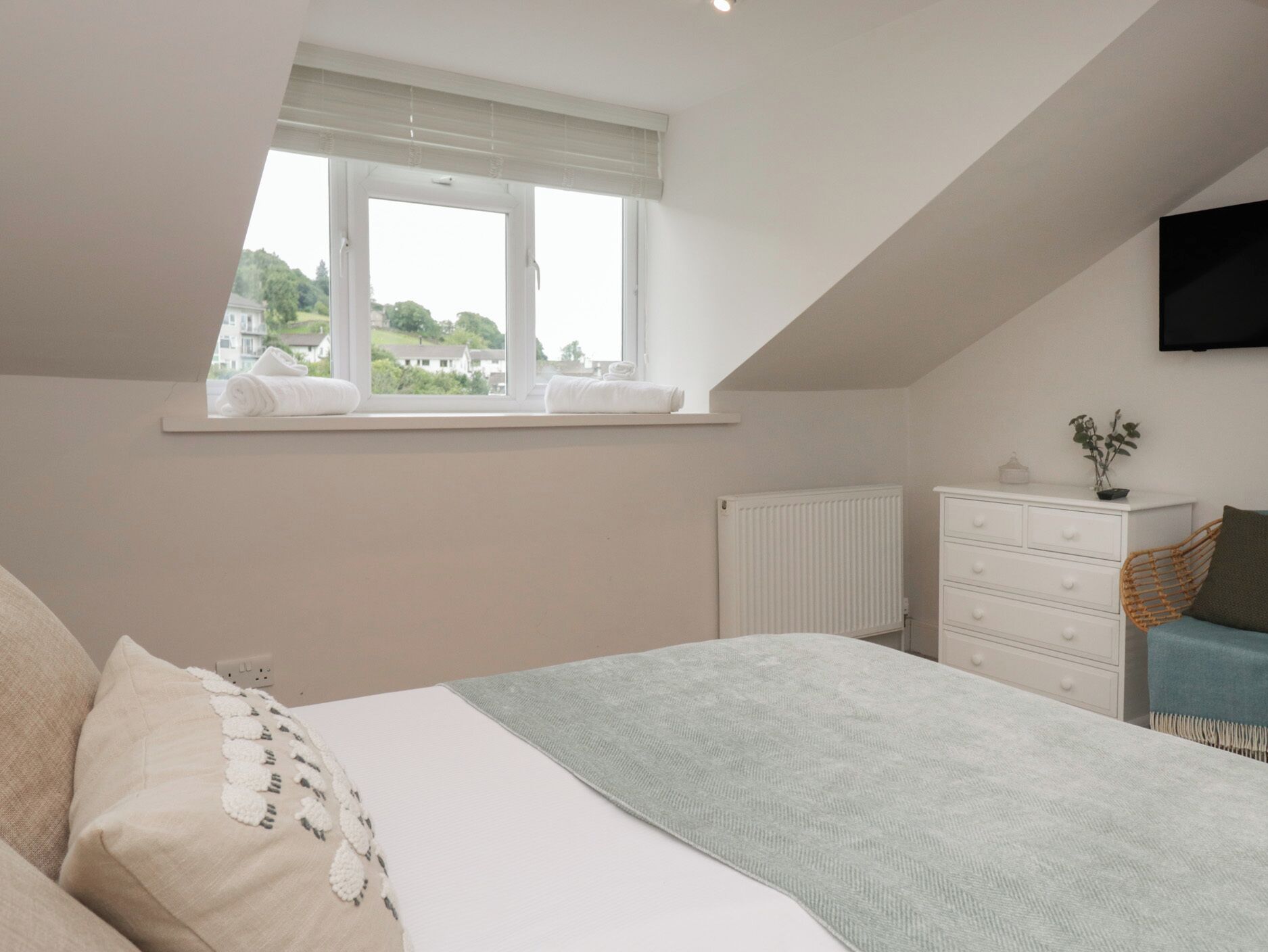 A bedroom with a large bed, a window with white blinds, a radiator, a dresser, and a television mounted on the wall.
