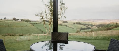 Ein runder Glastisch mit zwei Korbstühlen, draußen aufgestellt mit Blick auf sanfte Hügel und einen klaren Himmel.
