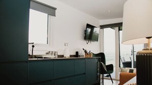 A modern kitchen with dark cabinetry, a flat-screen TV mounted above the counter, and a view of the outdoors through a large window. - Aganmore Farm Glamping (Ballycastle)