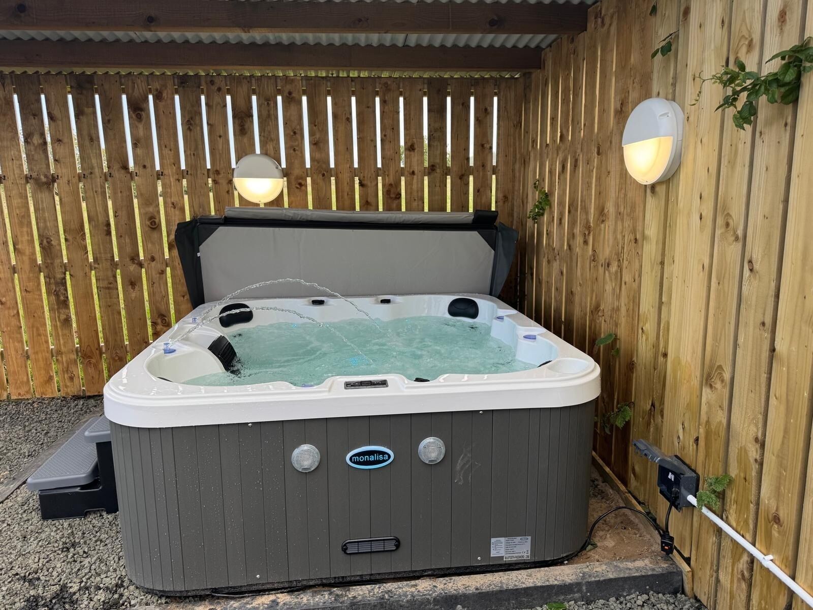 A hot tub with a wooden fence backdrop, a step for access, and a power outlet on the ground.