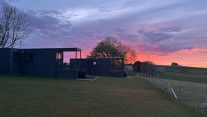 A modern building with a glass facade, a small outdoor seating area, and a view of a sunset over a grassy field. - Aganmore Farm Glamping (Ballycastle)