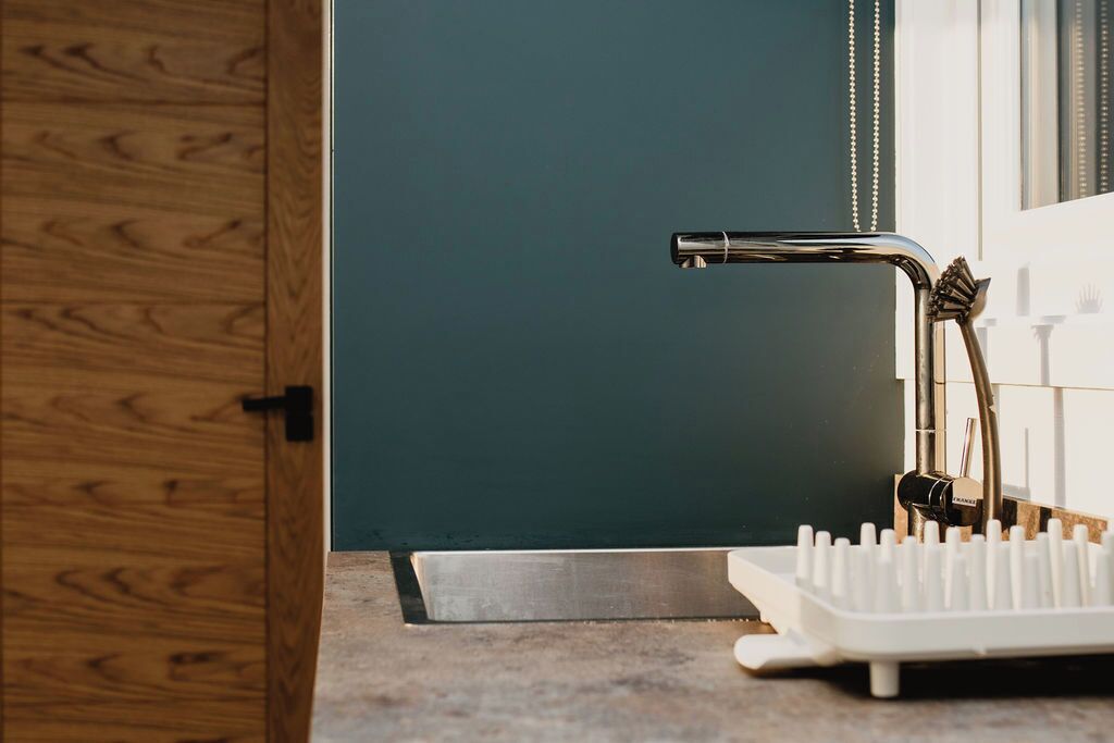 A bathroom with a wooden door, a dark teal wall, a chrome faucet, and a white toothbrush holder.