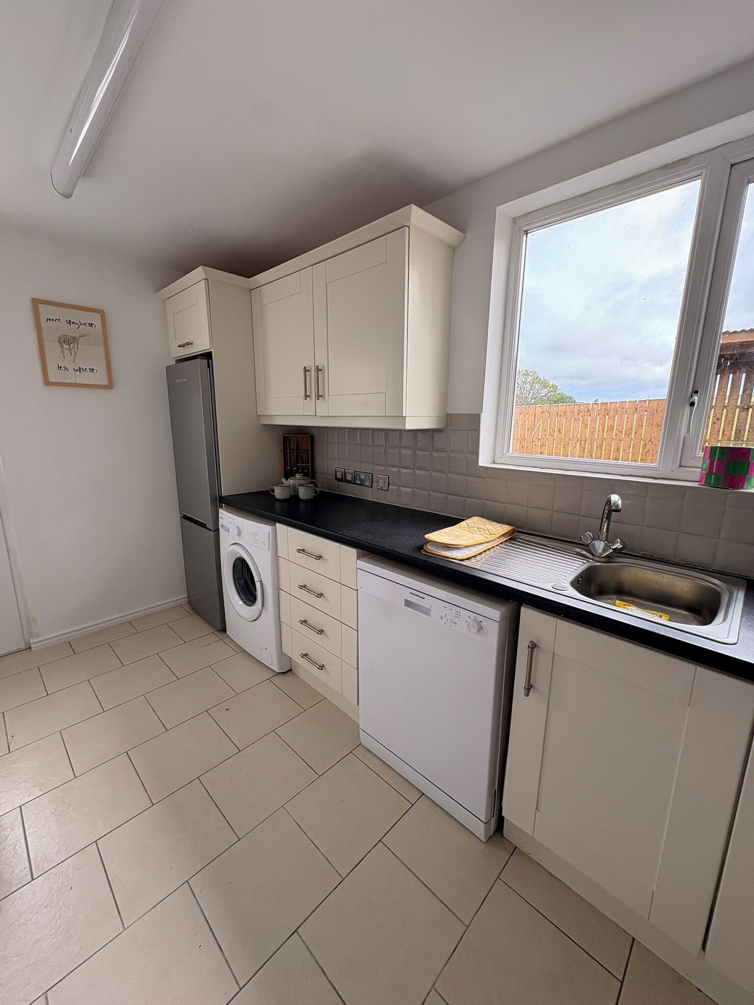 A compact kitchen with a washing machine, dishwasher, and a window with a view of a wooden fence.