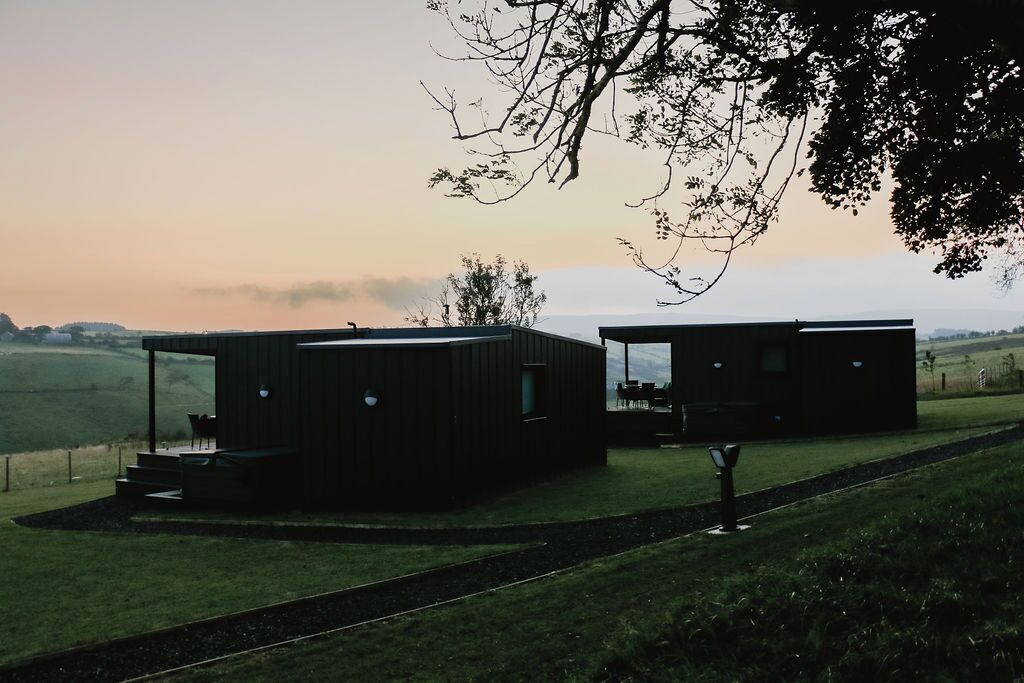 A modern, two-story building with a flat roof and large windows, situated on a grassy hill with a view of the countryside.