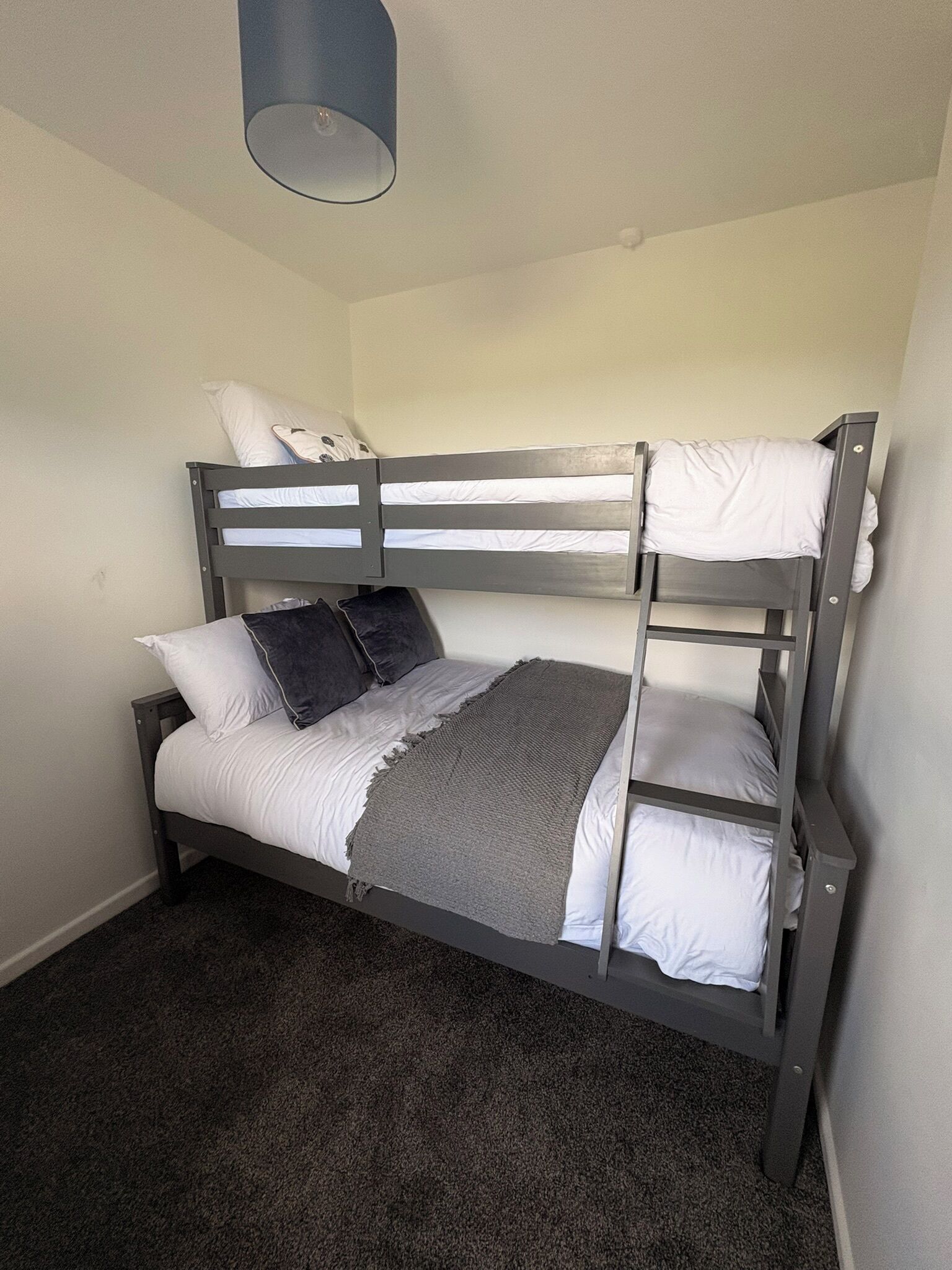 Een stapelbed met witte lakens en een grijze deken, een klein rond tafeltje en een lamp erboven.