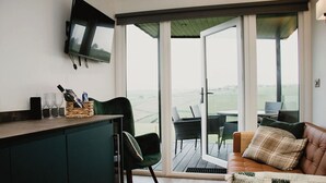 A modern living room with a television, a green chair, a brown sofa, and a view of the outdoors through large sliding doors. - Aganmore Farm Glamping (Ballycastle)