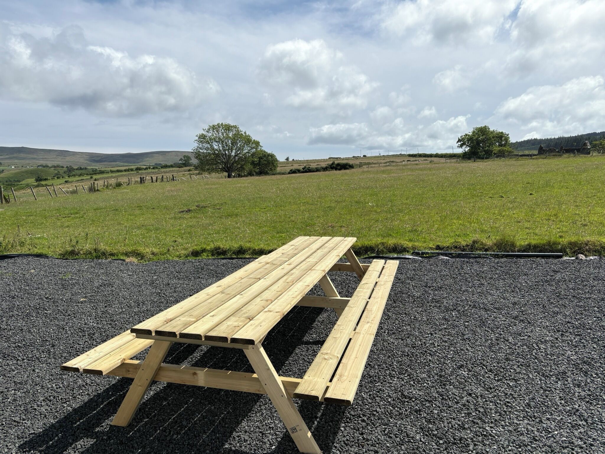 Une table de pique-nique en bois, posée sur une surface de gravier, avec un champ herbeux et des arbres en arrière-plan.