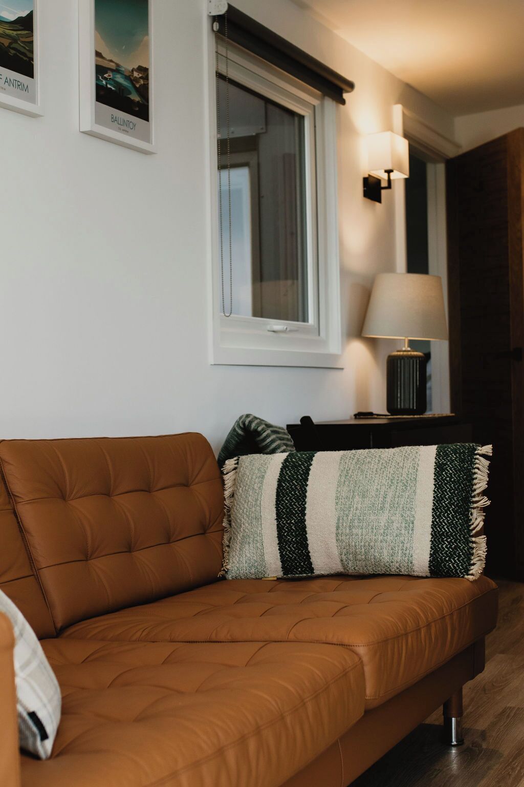 A brown leather sofa with a striped throw pillow, a window with curtains, a wall-mounted lamp, and framed artwork on the wall.