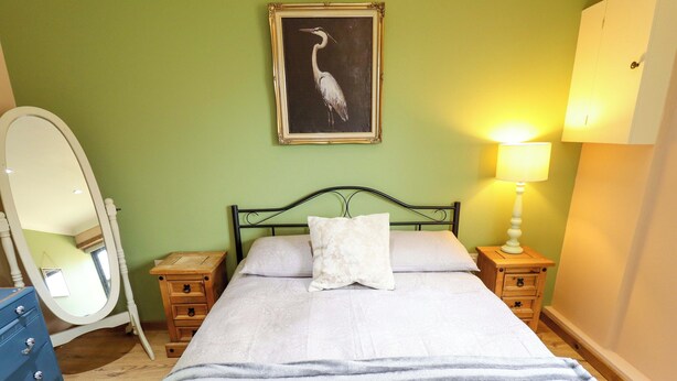 Een slaapkamer met een bed, nachtkastje, kaptafel, spiegel, lamp en een lijst met een afbeelding aan de muur.