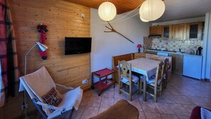 A cozy room with wooden walls, a TV mounted on the wall, a dining table with chairs, and a kitchen area with cabinets and a tiled backsplash.