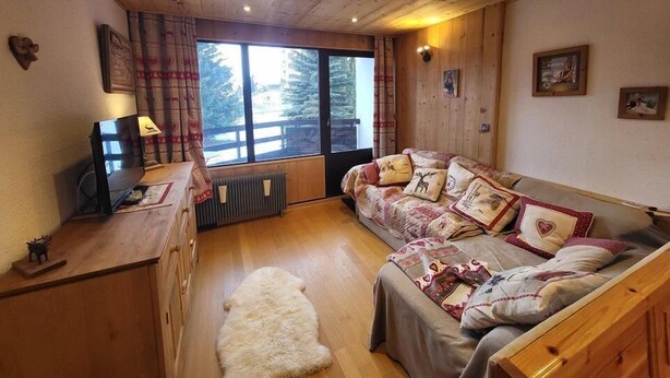Interior acogedor de una cabaña con techo de madera, un sofá con cojines estampados, una alfombra pequeña y una ventana con vistas a árboles.