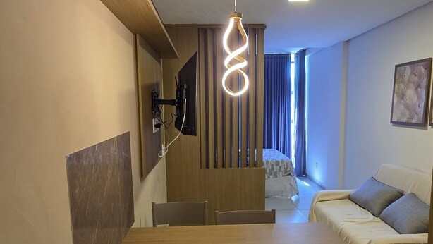 Une chambre d’hôtel compacte comprenant une table à manger en bois, deux chaises, une étagère intégrée, un téléviseur fixé au mur, un lit avec une tête de lit et une fenêtre avec des rideaux.