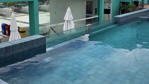 Une piscine dont les bords sont recouverts de carreaux, séparée du reste par une barrière en verre, et ombragée par des parasols blancs.