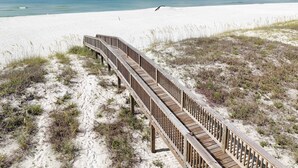 Ein hölzerner Steg führt zu einem Strand mit Sand und spärlicher Vegetation.