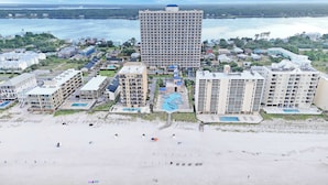 An aerial view of a coastal resort with multiple high-rise buildings, a sandy beach, and a large swimming pool.