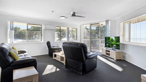 A spacious living room with a black leather sofa, a black leather chair, a wooden coffee table, and a TV on a wooden stand. Large windows provide ample natural light and a view of the outdoors. - Unit 13 Charlton on the Esplanade (Torquay)