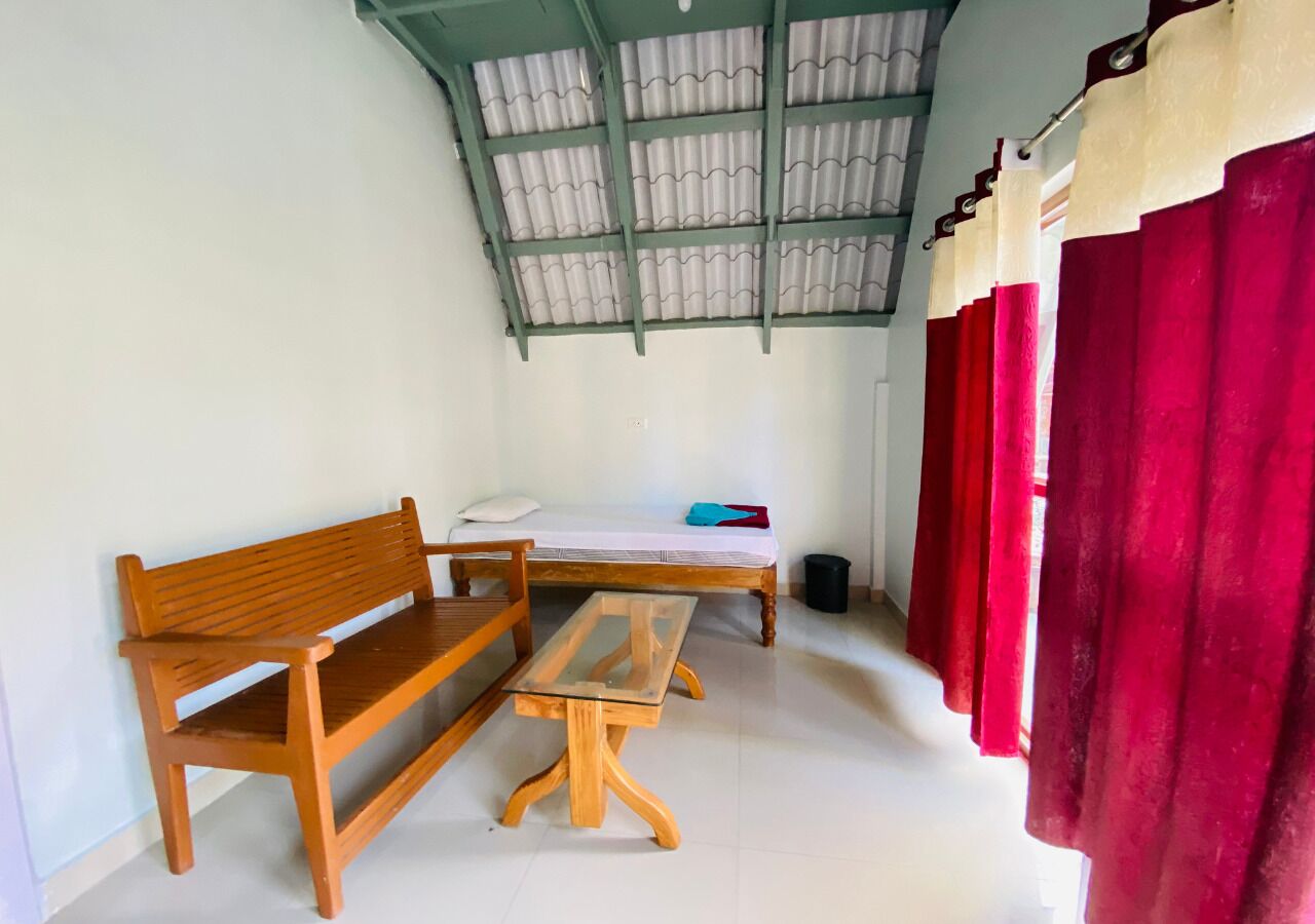 Um quarto com um banco de madeira, uma mesa pequena, uma cama com lençol branco e uma cortina vermelha.
