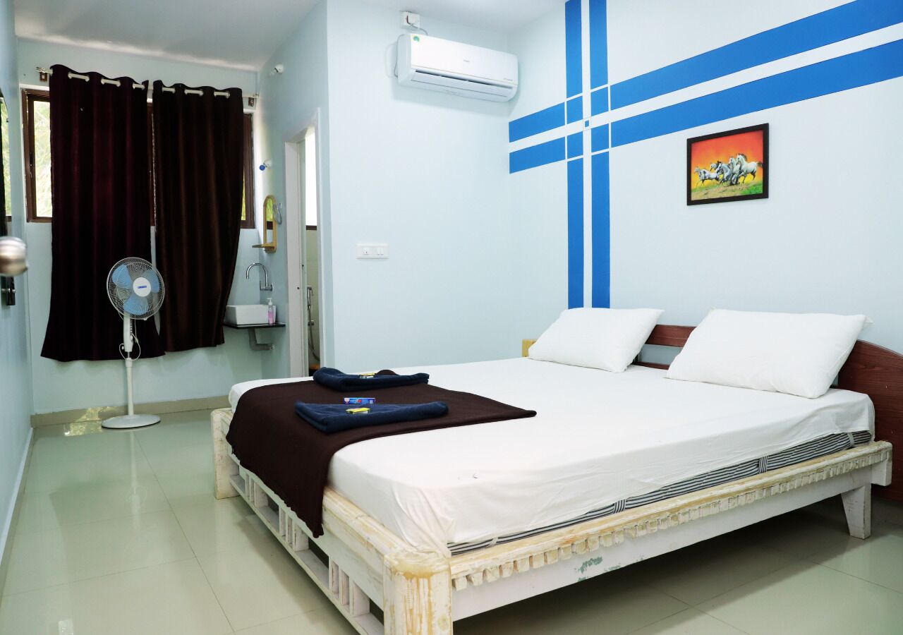 Una cama individual con ropa de cama blanca, un ventilador de pie, un aire acondicionado instalado en la pared y un diseño de pared a rayas azul y blanco.