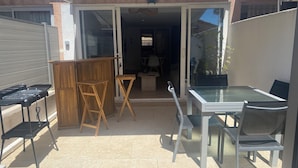 Um pátio externo com mesa e cadeiras de vidro, uma mesa dobrável de madeira e uma churrasqueira.