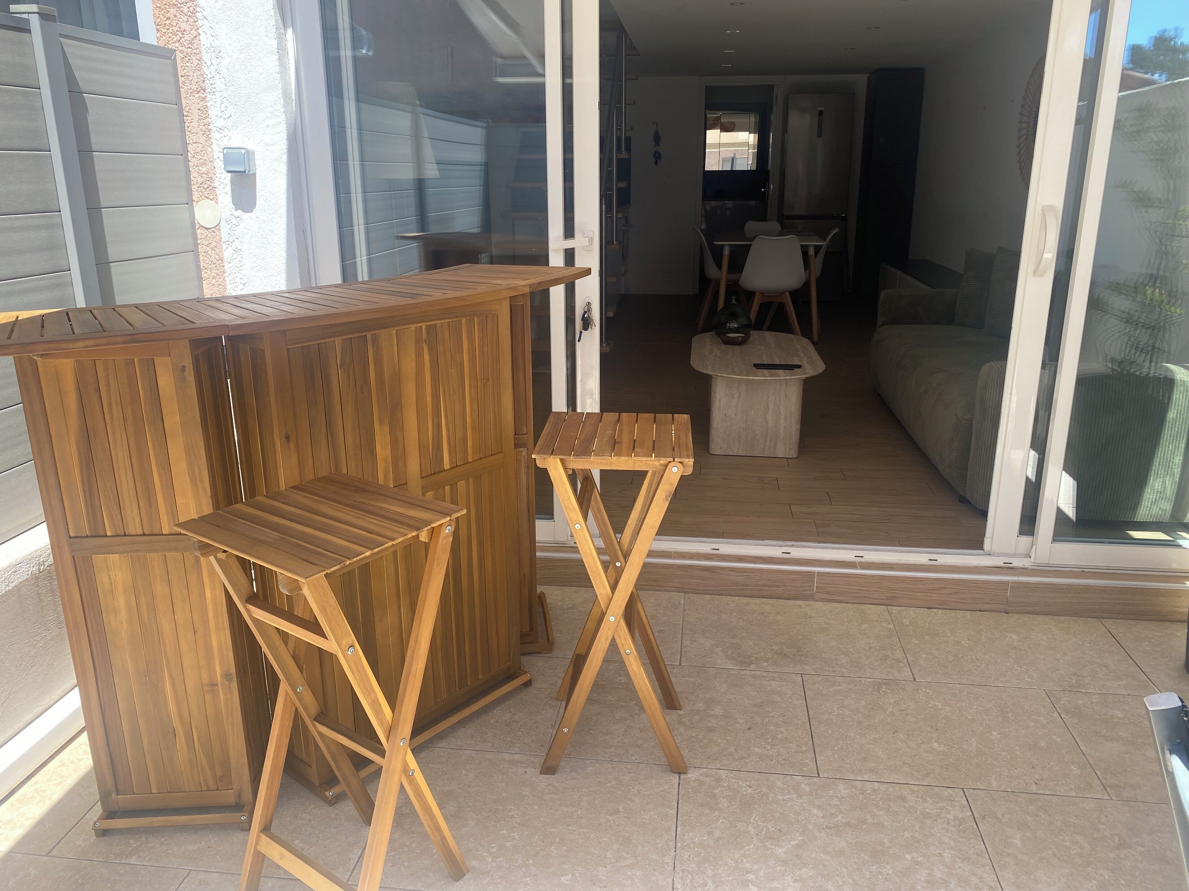 Un área de estar exterior de madera con una mesa y sillas, una mesita redonda pequeña y un ventanal corredero que da a un espacio interior con un sofá y una mesa de comedor.