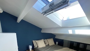Quarto com claraboia, cama com colcha bege, mesa de cabeceira com abajur e guarda-roupa branco.
