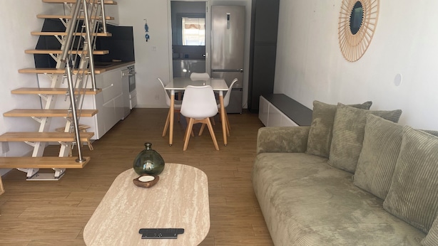 Uma sala de estar moderna com escada de madeira, um sofá, uma mesa de centro com controle remoto e uma área de cozinha ao fundo.