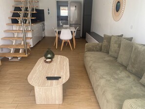 A modern living room with a wooden staircase, a sofa, a coffee table with a remote control, and a kitchen area in the background. - Villa Club 4/5 persons in secured residence with swimming pool, 400 m from the beach (Sanary-sur-Mer)