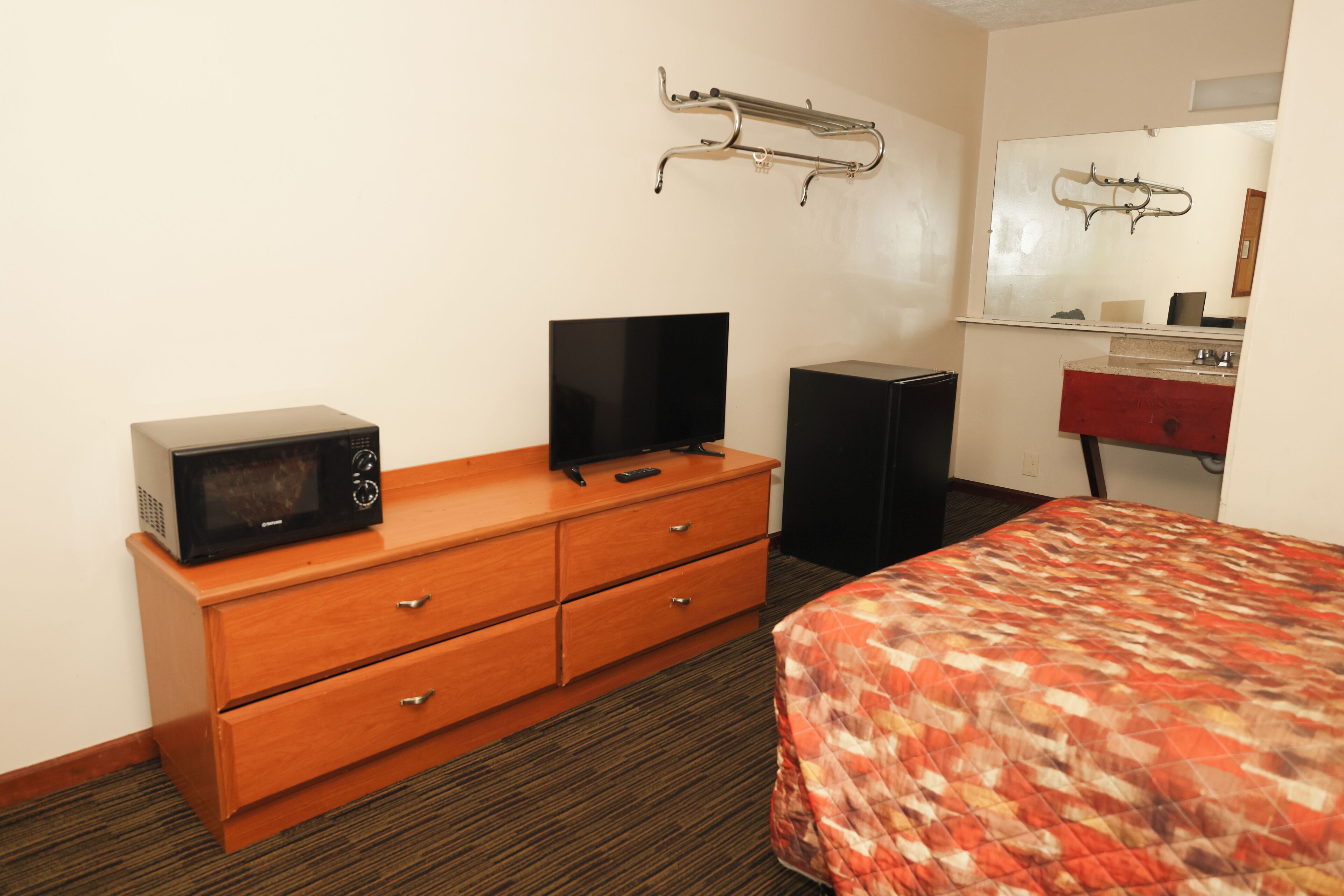 Habitación de hotel con cómoda de madera, televisor de pantalla plana, microondas, altavoz y cama con colcha estampada.