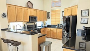 A modern kitchen with wooden cabinets, a stainless steel refrigerator, a microwave, and a stove. There are two bar stools and a trash can. - NEW Modern 3bd Home in Palm Springs,PGA Country Club the Ultimate Retreat (Cathedral City)