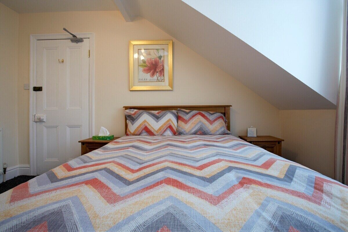 A bedroom with a chevron-patterned bedspread, a wooden headboard, a framed floral artwork on the wall, a white door, and a small bedside table with a lamp.
