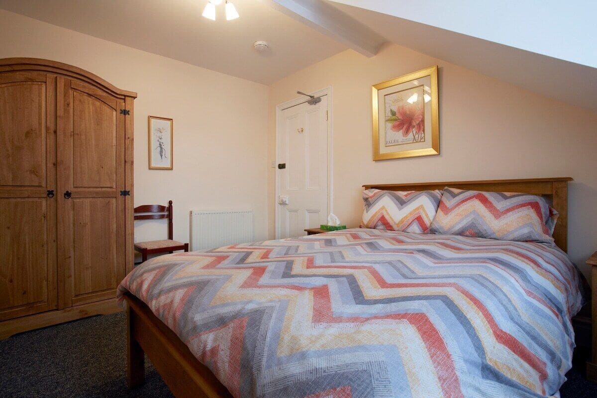 A bedroom with a bed, a wooden wardrobe, a chair, a framed picture, and a door.