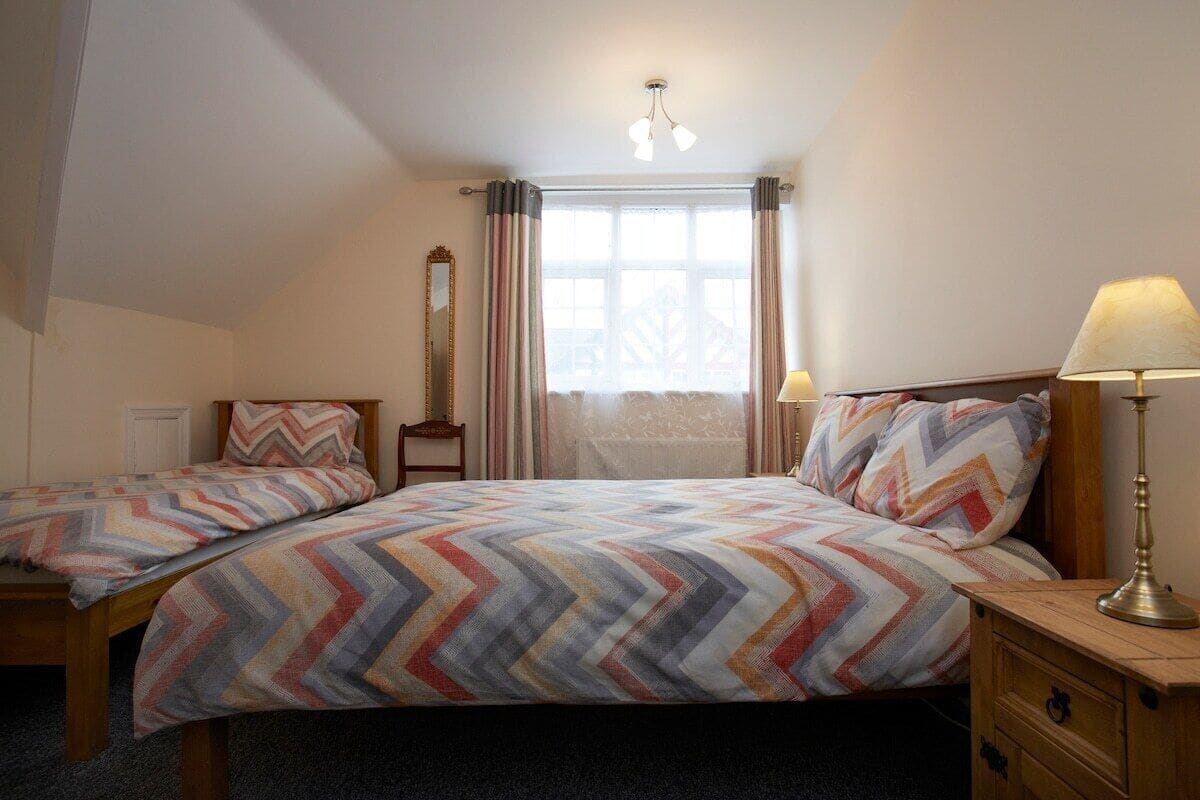 A bedroom with two beds, a window with curtains, a mirror, and a bedside lamp.
