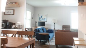 A modern living room with a blue sofa, a wooden dining table, and a coffee bar area.