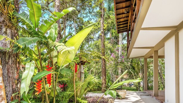 A covered walkway with a concrete path, surrounded by lush tropical vegetation and a wooden structure. - Villaggio Dei Pini (Ubatuba)