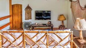 A room with a wooden ceiling, a TV mounted on the wall, a wooden door, a table with a lamp, and a woven chair. - Villaggio Dei Pini (Ubatuba)