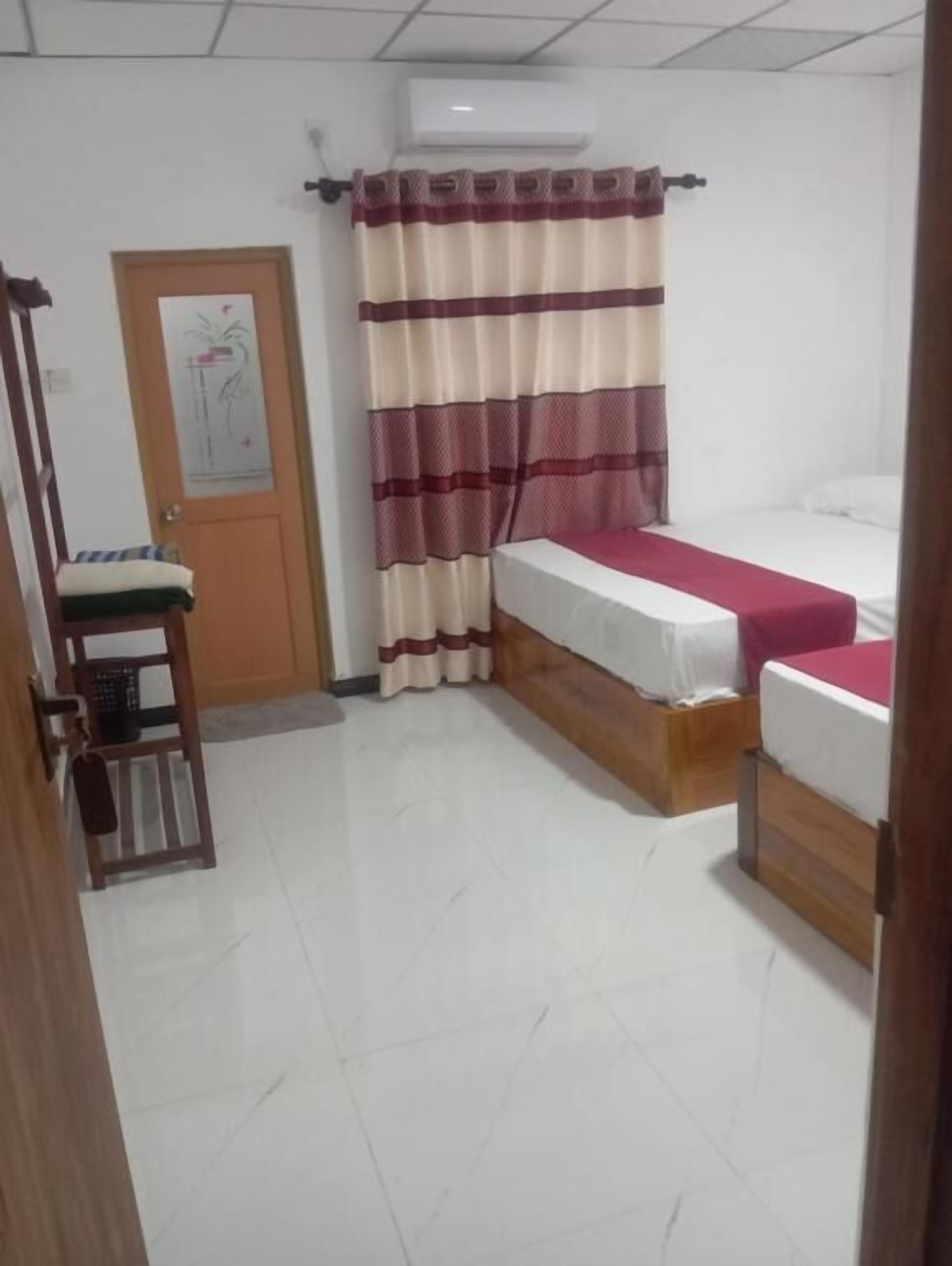 Una habitación pequeña y limpia con dos camas, una silla de madera, una puerta con un cartel y una ventana con cortinas.