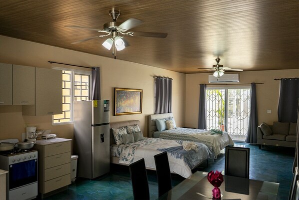 A compact hotel room with a kitchenette, a bed with a floral bedspread, a ceiling fan, and a view through the window.
