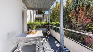 Un balcón con mesa y sillas, una planta en maceta y vistas a un jardín y edificios cercanos.