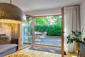 Een moderne woonkamer met een bank, een eettafel en stoelen, een groot raam met uitzicht op groen, en een hanglamp.