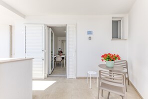 An open door leading to a dining area with a table and chairs, a small stool, and a potted plant with red flowers.